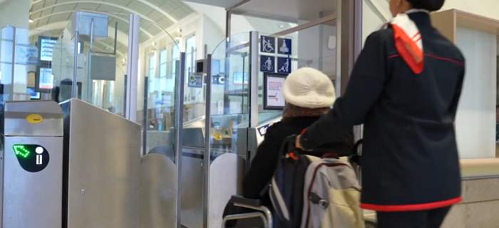 Procédure Assist'enGare : un agent SNCF prend en charge une voyageuse à mobilité réduite qui se déplace en fauteuil roulant. Elles franchissent les portiques de contrôle élargis permettant d'accéder à l'intérieur de la gare.
